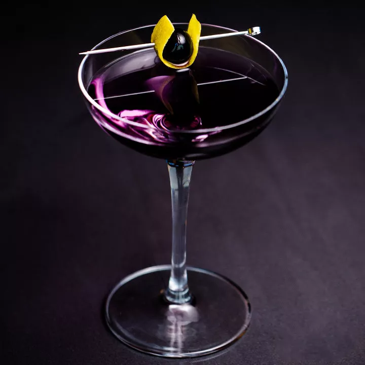 一个优雅的轿跑车玻璃杯里盛着深紫罗兰色的饮料，上面装饰着用柠檬皮包裹的樱桃，用尖头刺穿。 玻璃位于深灰色的表面上。